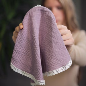 May include: A soft, purple muslin baby burp cloth with white lace trim. The cloth is folded in half and held by a person's hands.