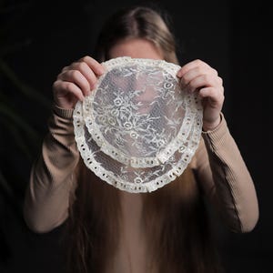 May include: A white lace doily with a scalloped edge. The doily is being held in front of a person's face.