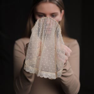May include: A cream-coloured veil with a dotted pattern and a scalloped lace trim. The veil is held up, showcasing its intricate design and texture. The background is a dark, solid colour.