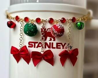 May include: A white mug with a gold chain and red bows hanging from it. The chain has red beads, green and red ornaments, and gold bells. The mug has the word "STANLEY" printed on it.