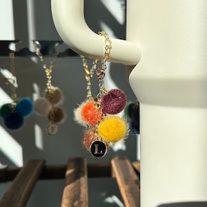 May include: A gold chain with a black letter "L" charm and five colourful fuzzy pom-poms attached. The pom-poms are orange, red, yellow, brown, and white.