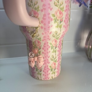 May include: A pink and gold charm with a pink bow and a letter K charm. The charm is attached to a gold chain and is hanging from a pink and white floral tumbler.