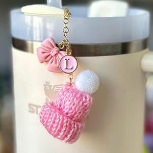 May include: A pink knit hat charm with a white pom pom and a pink bow with a gold chain and a pink letter L charm.