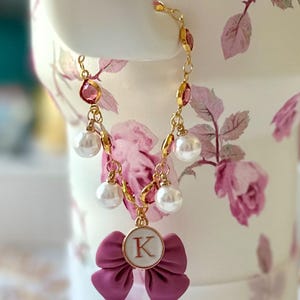 May include: A gold chain necklace with pink and white beads, a pink bow charm with a gold letter K, and pearl accents.