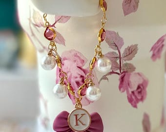 May include: A gold chain necklace with pink and white beads, a pink bow charm with a gold letter K, and pearl accents.