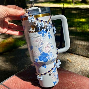 May include: A light blue tumbler with a handle, decorated with blue and white floral patterns and the text "LOVESHACKFANCY". The tumbler is adorned with a gold chain, blue bows, and pearls. The tumbler is held by a person.