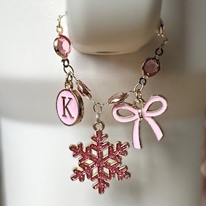 May include: A pink and gold charm bracelet featuring a snowflake, bow, and initial "K" pendant. The bracelet has pink gemstone accents and is suspended against a white background. The charms are detailed with gold trim.