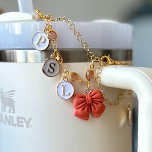 May include: A gold-toned chain charm accessory featuring three circular white enamel charms with the letters P, S, and L in gold, interspersed with small amber-colored beads and a prominent coral-colored bow charm attached to a metal clasp.