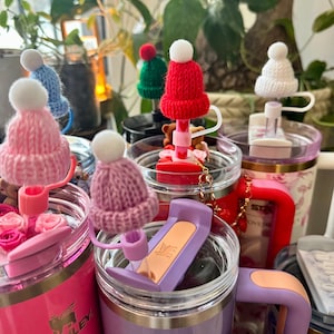May include: A collection of Stanley tumblers in various colors, each featuring a straw topper shaped like a miniature knitted beanie hat with a white pom-pom. The tumblers are pink, purple, and red, with some displaying floral patterns and bear charms.