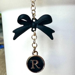 May include: A gold chain with a black bow and a round charm with the letter R in gold on a black background.