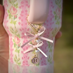 May include: A pink ceramic mug with a floral pattern and a gold chain with pink ribbon bows and pearl accents. The chain has a gold charm with the letter "H" on it.