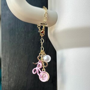 May include: A gold chain charm with a pink enamel letter "P" charm, a pink bow charm, and a white pearl charm.