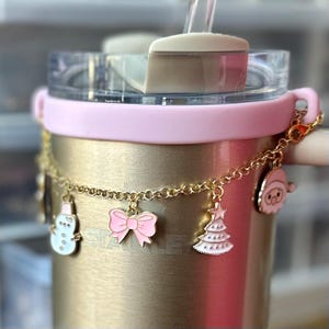 May include: A gold stainless steel tumbler with a pink silicone lid and a gold chain with charms. The charms include a snowman, a pink bow, a Christmas tree, and a cartoon character.