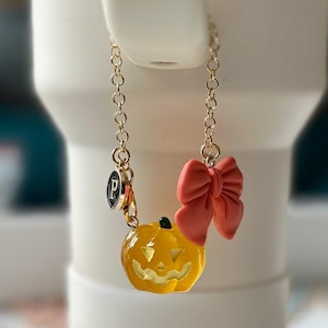 May include: A gold-toned metal chain charm featuring a translucent yellow jack-o'-lantern pumpkin with a green stem, a solid coral-coloured bow, and a small circular black and silver charm with the letter P engraved on its surface.