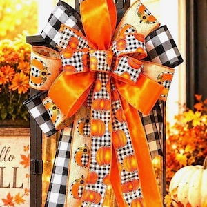 May include: A decorative bow featuring a mix of solid orange satin ribbon, black and white buffalo check ribbon, and a cream-colored ribbon printed with orange pumpkins and sunflowers. The bow has multiple loops and long, trailing ribbon tails.