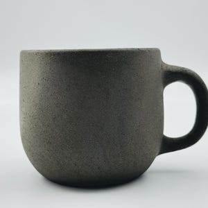 Puede incluir: Una taza de cerámica gris oscuro con cuerpo redondeado y asa. La taza tiene una superficie texturizada.