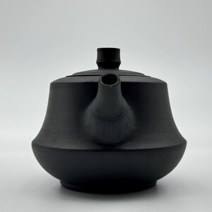 Puede incluir: Tetera de cerámica negra mate con cuerpo redondeado y boquilla corta. La tetera tiene tapa y asa en la parte superior. El diseño es minimalista y moderno, adecuado para preparar té.