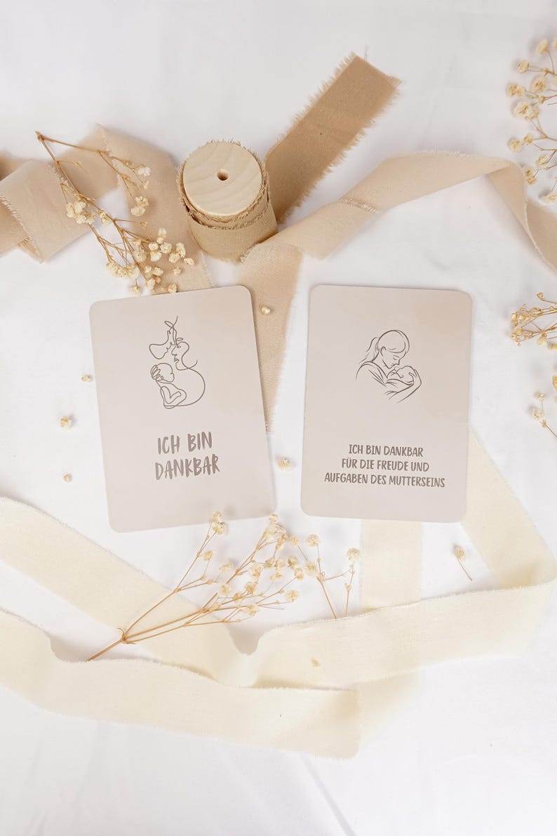 May include: Two rectangular beige cards featuring black line art illustrations of a parent holding a baby. The left card displays the text ICH BIN DANKBAR. The right card displays the text ICH BIN DANKBAR F&Uuml;R DIE FREUDE UND AUFGABEN DES MUTTERSEINS.