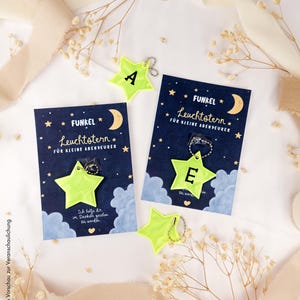 Könnte beinhalten: Neon-gelbe sternförmige Reflektoren mit den schwarzen Buchstaben A und E, befestigt an silbernen Kugelkettchen. Jeder Reflektor ist auf einer dunkelblauen Karte mit Mond, Sternen, Wolken und dem Text Funkel Leuchtstern für kleine Abenteurer präsentiert.