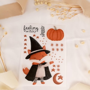 Könnte beinhalten: Ein Aufkleberbogen mit einem Fuchs, der einen schwarzen Hexenhut und Umhang, gestreifte Leggings und einen Kürbis trägt. Der Text lautet feeling witchy und spooky season. Weitere Elemente sind kleine Sterne, ein Halbmond und verschiedene Sternformen.