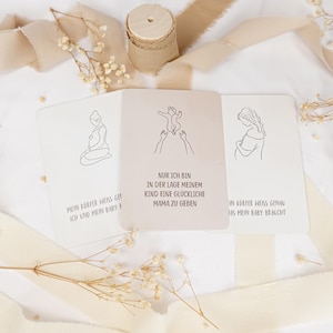 May include: Three rectangular cards with line art illustrations and German text. The center card shows a baby being held up with the text 'NUR ICH BIN IN DER LAGE MEINEM KIND EINE GL&Uuml;CKLICHE MAMA ZU GEBEN'. The cards are surrounded by beige ribbon and dried flowers.