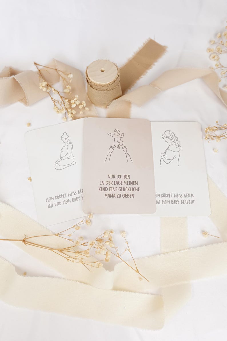 May include: Three rectangular cards with line art illustrations and German text. The center card shows a baby being held up with the text 'NUR ICH BIN IN DER LAGE MEINEM KIND EINE GL&Uuml;CKLICHE MAMA ZU GEBEN'. The cards are surrounded by beige ribbon and dried flowers.