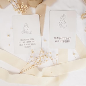 May include: Two rectangular off-white cards featuring black line art illustrations of pregnant figures. The left card reads: MEIN K&Ouml;RPER IST IN DER LAGE, MEINEM BABY ALLES ZU GEBEN WAS ES BRAUCHT. The right card reads: MEIN K&Ouml;RPER DARF SICH VER&Auml;NDERN.