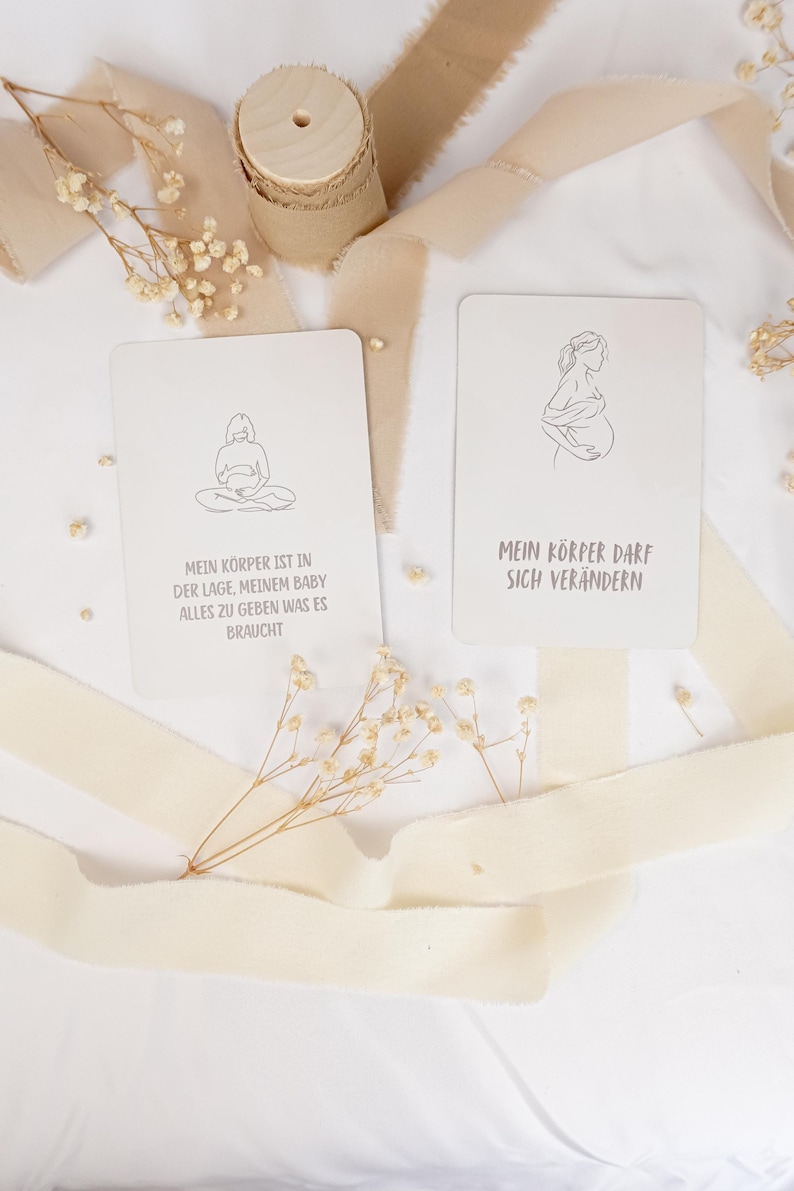 May include: Two rectangular off-white cards featuring black line art illustrations of pregnant figures. The left card reads: MEIN K&Ouml;RPER IST IN DER LAGE, MEINEM BABY ALLES ZU GEBEN WAS ES BRAUCHT. The right card reads: MEIN K&Ouml;RPER DARF SICH VER&Auml;NDERN.