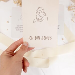 May include: A rectangular off-white card featuring a minimalist line drawing of a person holding a baby. Below the illustration, the text ICH BIN GENUG is printed in a dark gray sans-serif font. The card has rounded corners and a smooth matte finish.