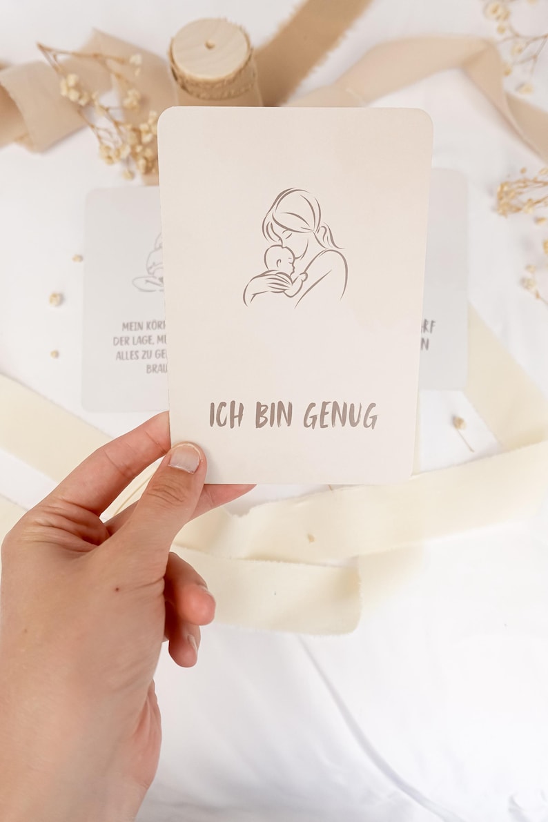 May include: A rectangular off-white card featuring a minimalist line drawing of a person holding a baby. Below the illustration, the text ICH BIN GENUG is printed in a dark gray sans-serif font. The card has rounded corners and a smooth matte finish.