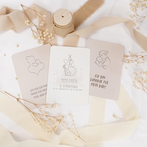 May include: Three rectangular cards with line art illustrations of a parent and baby, featuring German text including "MAMA KARTEN" and "ICH BIN DANKBAR F&Uuml;R MEIN BABY." The cards are arranged with a wooden spool and beige ribbon on a white surface.