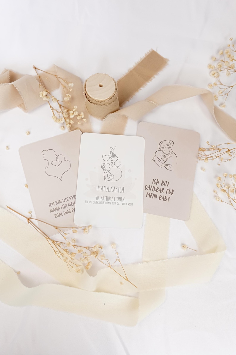 May include: Three rectangular cards with line art illustrations of a parent and baby, featuring German text including "MAMA KARTEN" and "ICH BIN DANKBAR F&Uuml;R MEIN BABY." The cards are arranged with a wooden spool and beige ribbon on a white surface.
