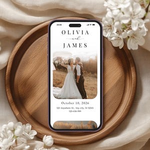 May include: A smartphone displays a wedding invitation with the names "Olivia and James" above a photo of a couple. The phone rests on a wooden tray, surrounded by white flowers and a beige fabric.