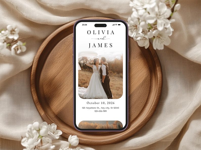 May include: A smartphone displays a wedding invitation with the names "Olivia and James" above a photo of a couple. The phone rests on a wooden tray, surrounded by white flowers and a beige fabric.
