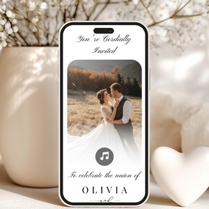 May include: A smartphone displays a wedding invitation with a photo of a couple embracing. The text reads "You're Cordially Invited" and "To celebrate the union of OLIVIA and". A white heart and vase with flowers are in the background.