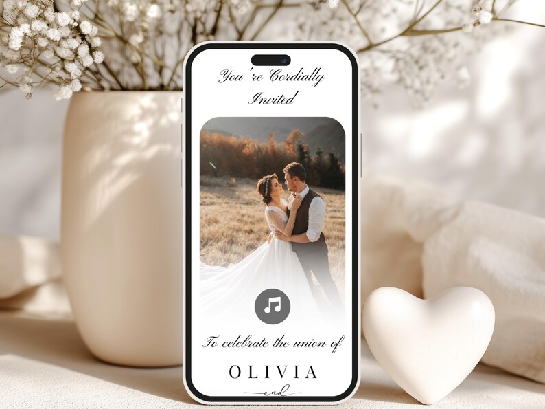 May include: A smartphone displays a wedding invitation with a photo of a couple embracing. The text reads "You're Cordially Invited" and "To celebrate the union of OLIVIA and". A white heart and vase with flowers are in the background.