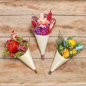 May include: Three dried flower bouquets in cream-colored paper cones. The bouquets feature a variety of colorful flowers, including red, purple, yellow, and pink blooms. The bouquets are arranged on a wooden surface.