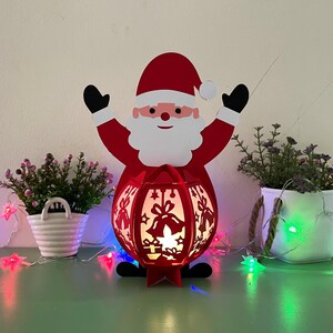 Op de afbeelding: Rode papieren kerstman-lantaarn met een witte baard en een brandende kaars erin. De lantaarn is versierd met bellen en sterren.