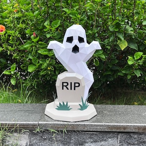 Peut inclure: Une figurine de fantôme blanc en style low-poly surplombant une pierre tombale gris clair avec les lettres RIP imprimées en noir. La pierre tombale repose sur une base gris clair assortie ornée de petits brins d'herbe en papier vert.