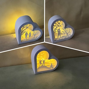 Peut inclure: Trois décorations lumineuses en papier en forme de cœur blanc avec des scènes différentes à l'intérieur. La première scène montre un couple se promenant dans une forêt, la deuxième montre un couple s'embrassant dans une forêt et la troisième montre un couple faisant de la randonnée dans un paysage de montagne.