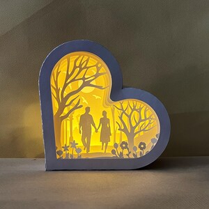 Peut inclure: Une lampe en papier en forme de cœur blanche avec une silhouette de couple se promenant dans une forêt. La lampe est éclairée de l'intérieur, projetant une lueur chaude.
