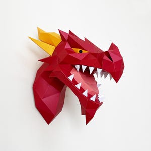 May include: A red papercraft dragon head with yellow horns and sharp white teeth. The dragon's mouth is open, revealing a pink interior. The artwork is mounted on a white wall, showcasing a geometric, polygonal design.