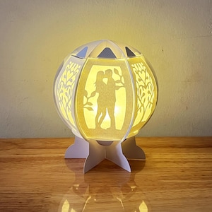 May include: A white paper lamp with a silhouette of a couple inside. The lamp has a round base and a white, star-shaped base.