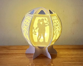 May include: A white paper lamp with a silhouette of a couple inside. The lamp has a round base and a white, star-shaped base.