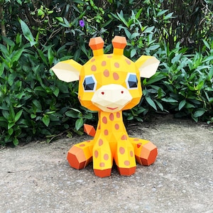 May include: A yellow papercraft giraffe with brown spots, big eyes, and a friendly smile. The giraffe is sitting on a grey surface.