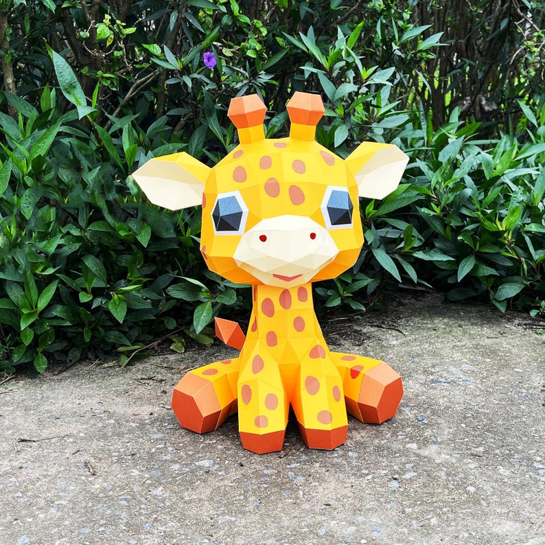 May include: A yellow papercraft giraffe with brown spots, big eyes, and a friendly smile. The giraffe is sitting on a grey surface.