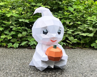 May include: A white geometric paper ghost figure holding an orange pumpkin. The ghost has black eyes and a red smile. The figure is sitting on a gray surface with green foliage in the background. A Halloween decoration.