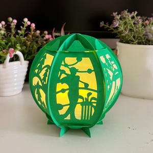 May include: A green paper lantern with a cut-out design of a golfer swinging a club. The lantern is illuminated from within, creating a warm glow.