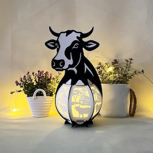 May include: A black and white silhouette of a cow's head with a white, intricately cut lampshade featuring a farm scene. The lampshade is illuminated with warm white light.