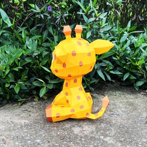 May include: A yellow papercraft giraffe with orange spots, sitting on a grey surface. The giraffe is looking to the right, with its head tilted slightly. The giraffe has a long neck and a long tail.